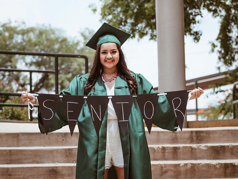 A charter school graduate wearing a green cap and gown holds a sign that says “senior” as she smiles for her post-graduation picture.